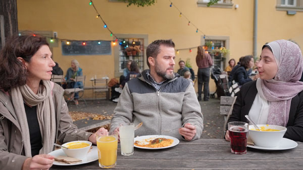 One man and two women having a conversation while they sit outside on a table at a cafe for lunch.