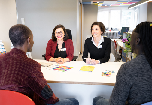 Two people sit across a table from two women offering consultation. The women are seated behind a white desk with brochures and papers in a modern office environment. 