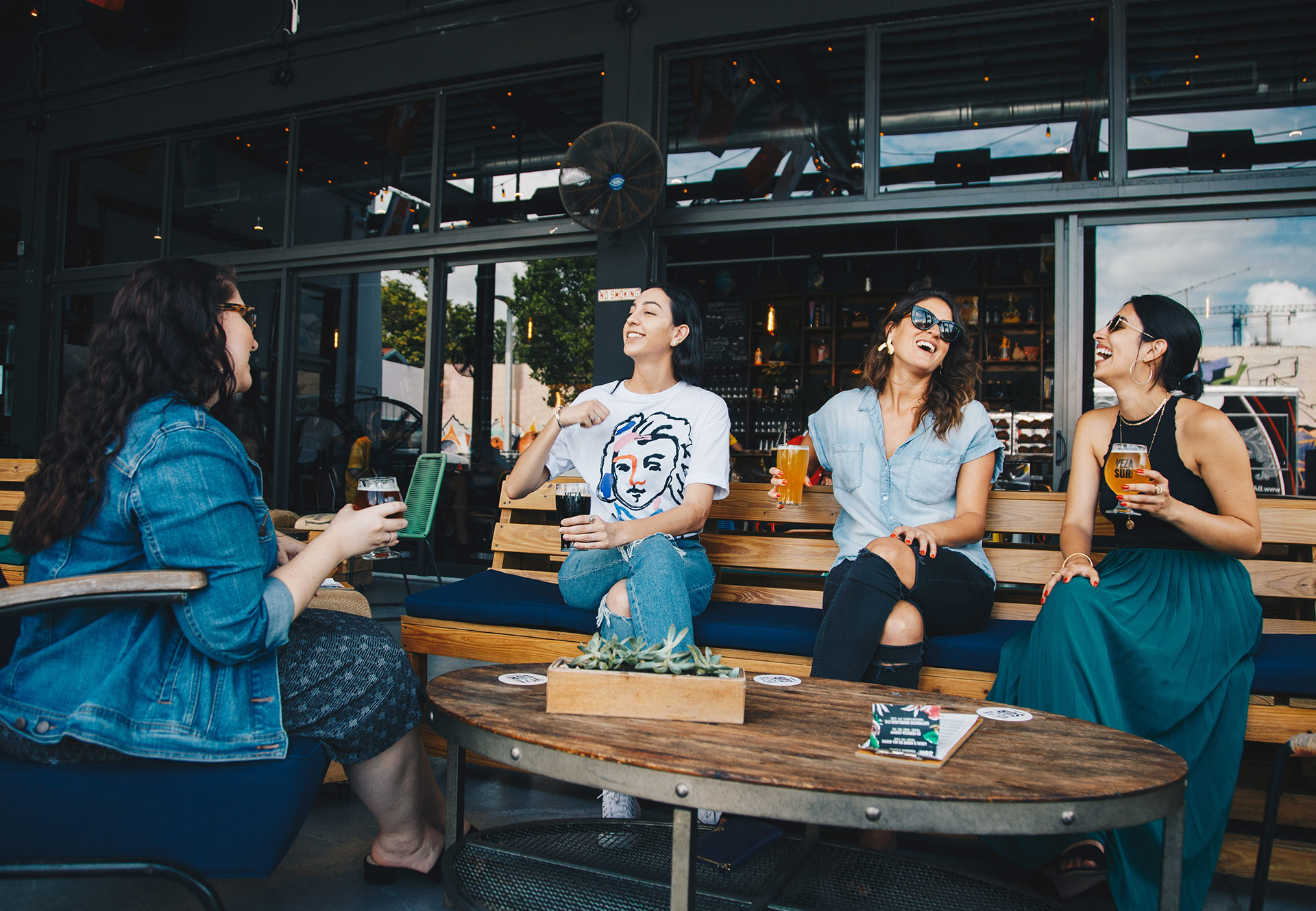 Four women are sitting on a bench at a table outside a pub, drinking beer and wine.