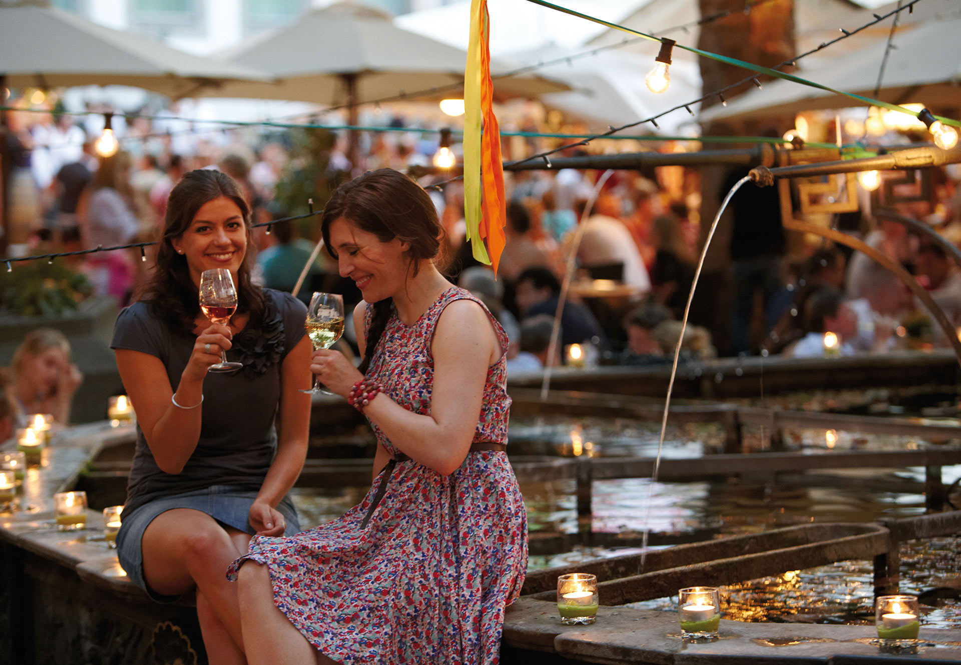 Two young women sit at the wine village fountain and each drink from a glass of wine.